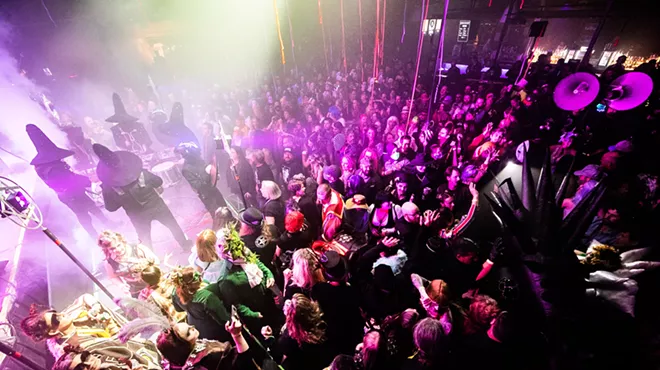 a crowd dances in a concert venue to masked drummers on stage wearing witch hats