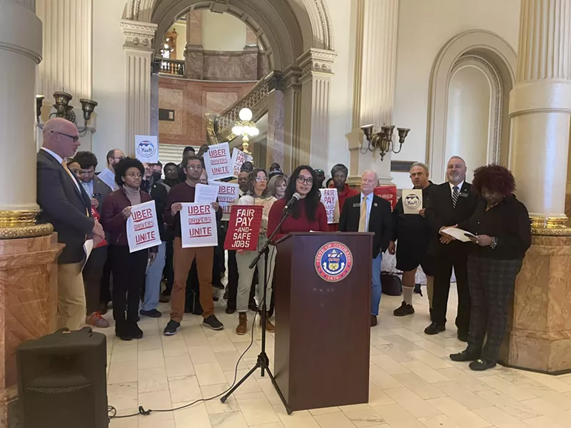 Image: A group of rideshare drivers and advocates stand behind a podium