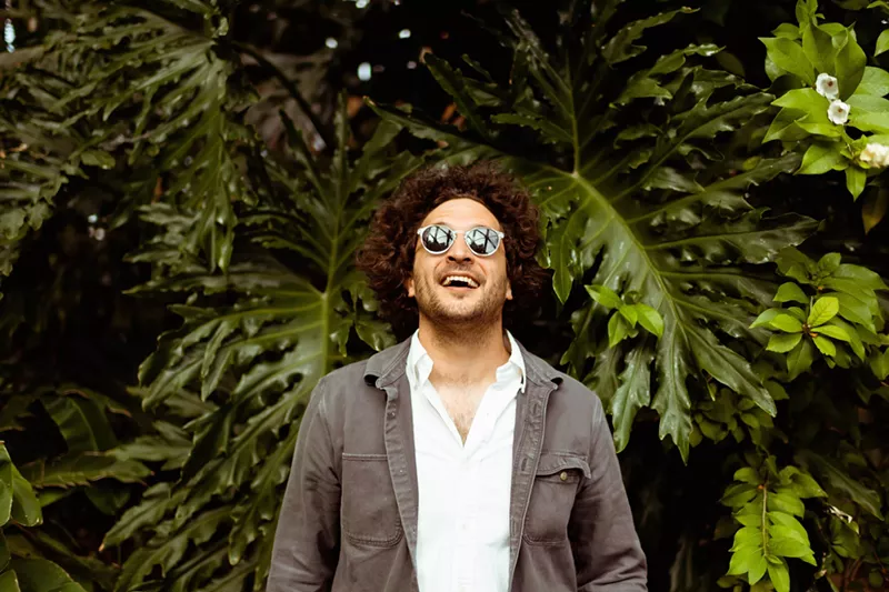 Image: man in sunglasses smiles in front of evergreen tree.