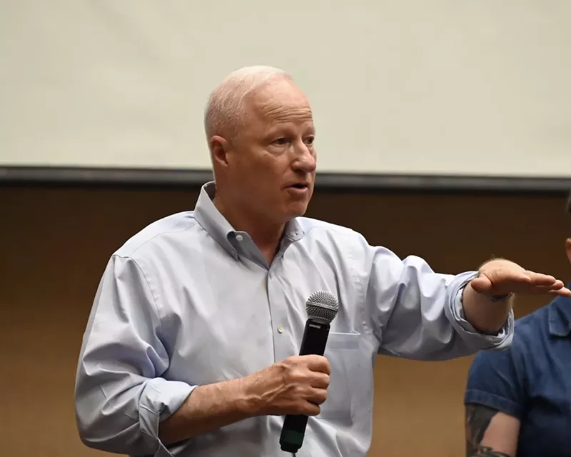 Image: Aurora Mayor Mike Coffman speaks with a microphone in his hand at a town hall.