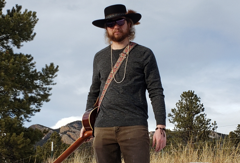 Image: Andrew Baker standing outside with guitar strapped around chest and shoulder.
