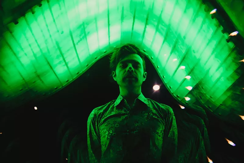 Image: a man stares seriously at the camera while cast in green lighting.