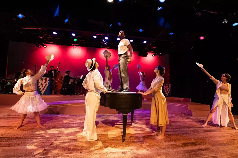 Image: Mykai Eastman stands atop a piano surrounded by the cast of Duke Ellington's Sophisticated Ladies