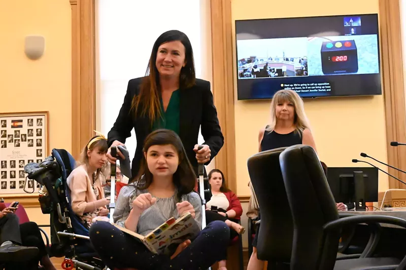 Image: Liz Cochran walks away from the panel after testify before the Senate Finance Committee about the importance of CBD products for her disabled daughter, who is in the wheelchair in front of her.