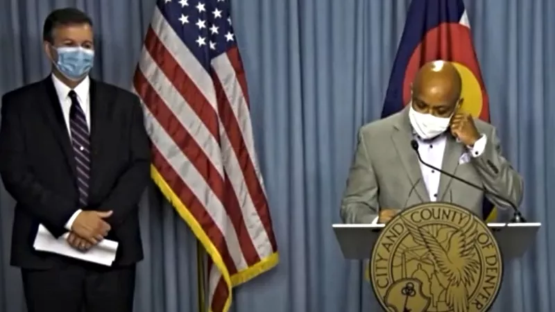 Image: Denver Mayor Michael Hancock, joined by Denver Public Health and Environment Executive Director Bob McDonald, removing his mask prior to speaking at a press event on July 15.