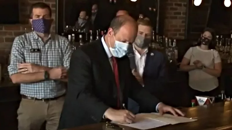 Image: A masked Governor Jared Polis on July 10, signing a bill to allow to-go liquor sales for pick-up and delivery.
