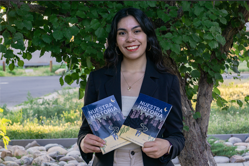 Image: A girl wearing a black blazer, gold hoop earrings and a bold red lip smiles while holding two books in front of greenery.