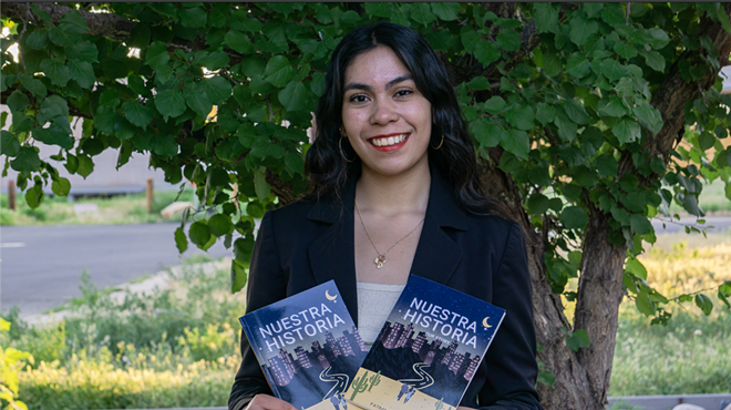 A girl wearing a black blazer, gold hoop earrings and a bold red lip smiles while holding two books in front of greenery.