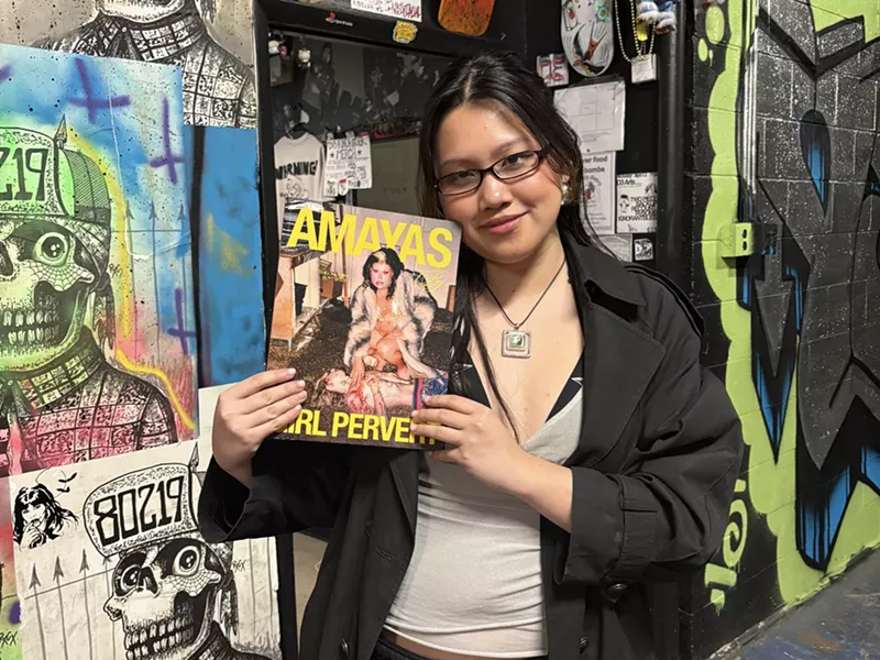 Image: Hà Pham stands in front of a mural wall while holding a copy of Girl Pervert: Volume One with cover star Amayas Gonzalez.
