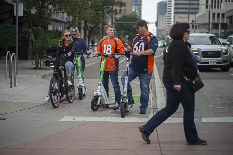 Image: A group of four adults riding standing e-scooters in downtown Denver.