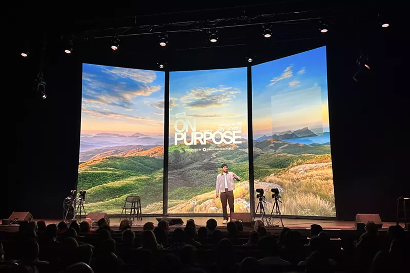 Image: Jay Shetty backdropped by a mountain scene on three digital panels while on stage at the Ellie Caulkins Opera House