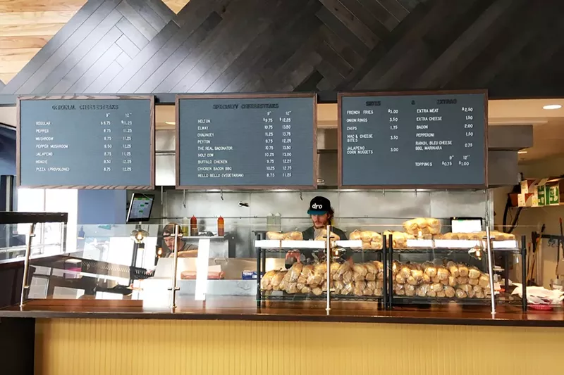 Image: a man in a hat standing behind a sandwich shop counter