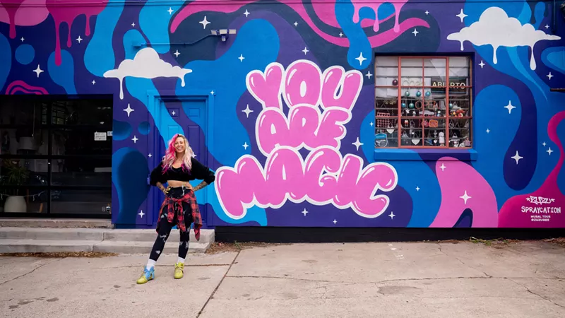Image: a woman stands in front of a colorful mural