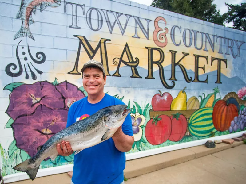 Image: a man holds a large salmon in front of a colorful mural with fruits and vegetables
