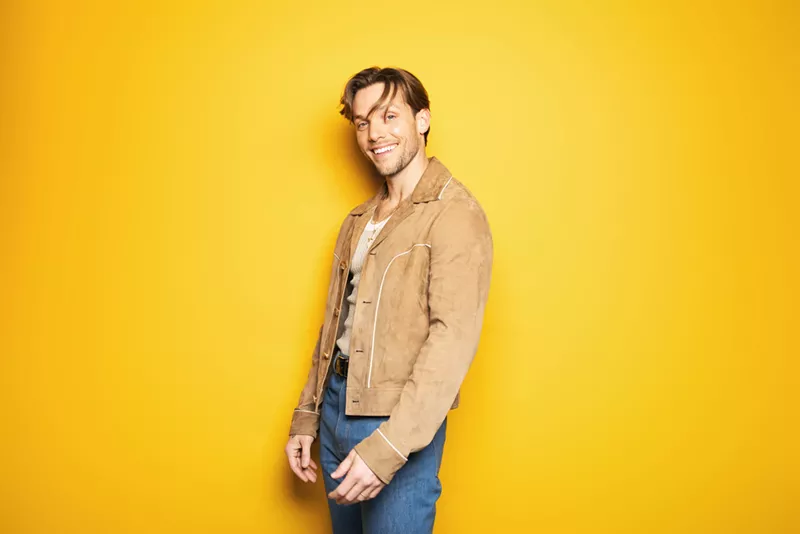 Image: Pop singer John K stands in front of a yellow background while wearing a brown jacket, blue jeans and a smile on his face.