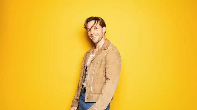 Pop singer John K stands in front of a yellow background while wearing a brown jacket, blue jeans and a smile on his face.