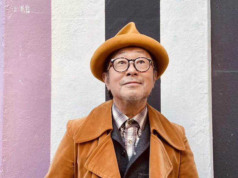 Image: Portrait of Senri Oe in brown hat and Jacket on vertically striped background.