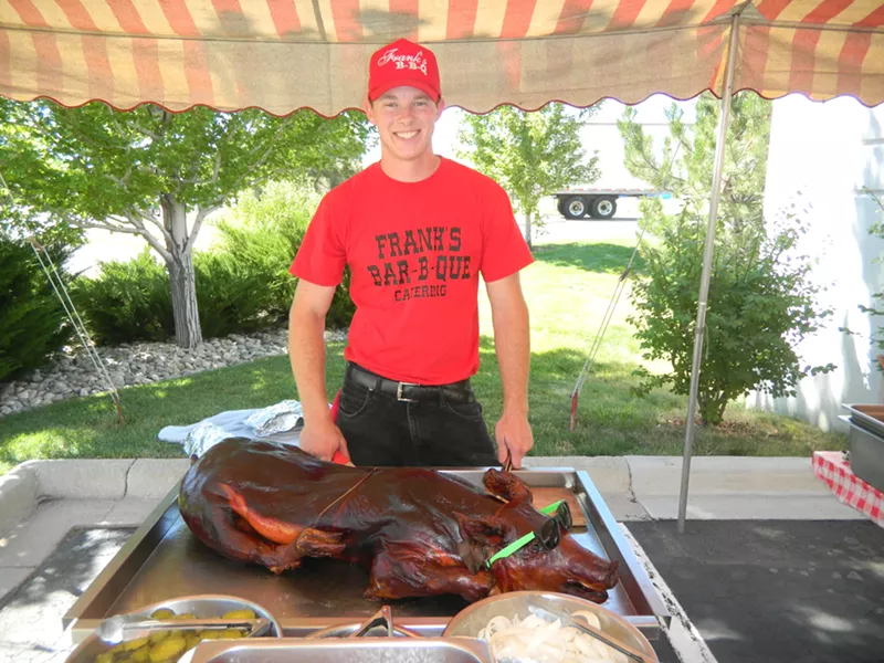 Image: a man in a red shirt standing in front of a whole barbecued pig