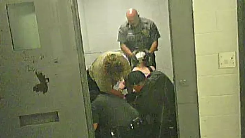 Image: In this screen capture from a video, a Fremont County deputy can be seen putting a restraint hold on a naked Carolyn O'Neal, her face covered with a spit mask, as other personnel try to force her foot back into a strap on a restraint chair.
