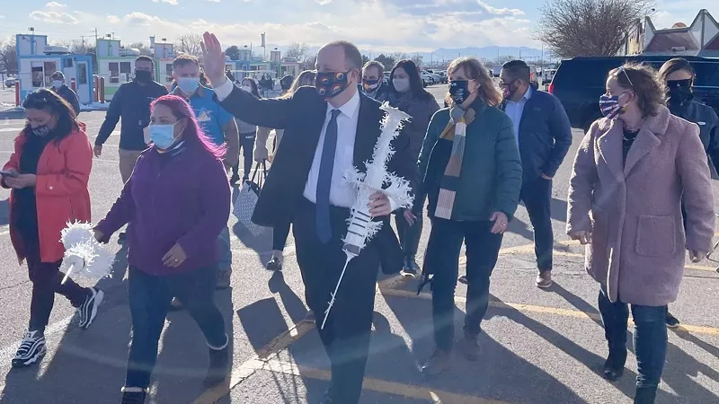 Image: Governor Jared Polis carrying a giant mock syringe during a visit to the Mile High Flea Market for a vaccination event.