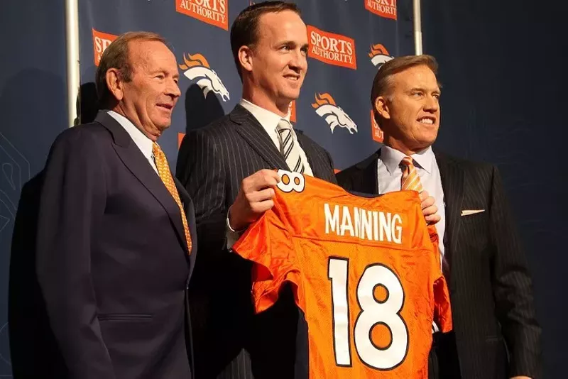 Image: Three men stand in a row, one holding up an orange jersey.