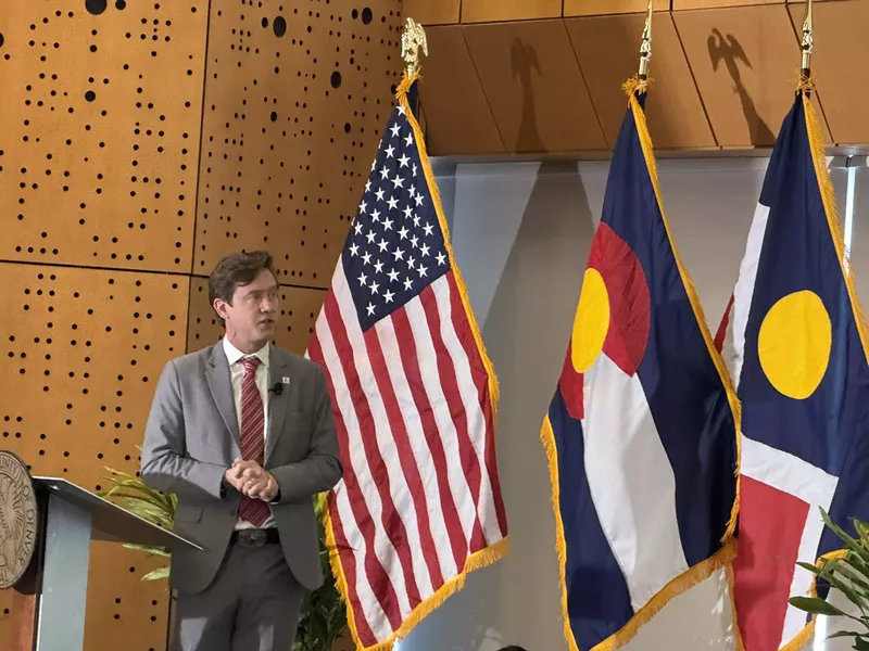 Image: Man in a suit speaks in front of American and Colorado flags