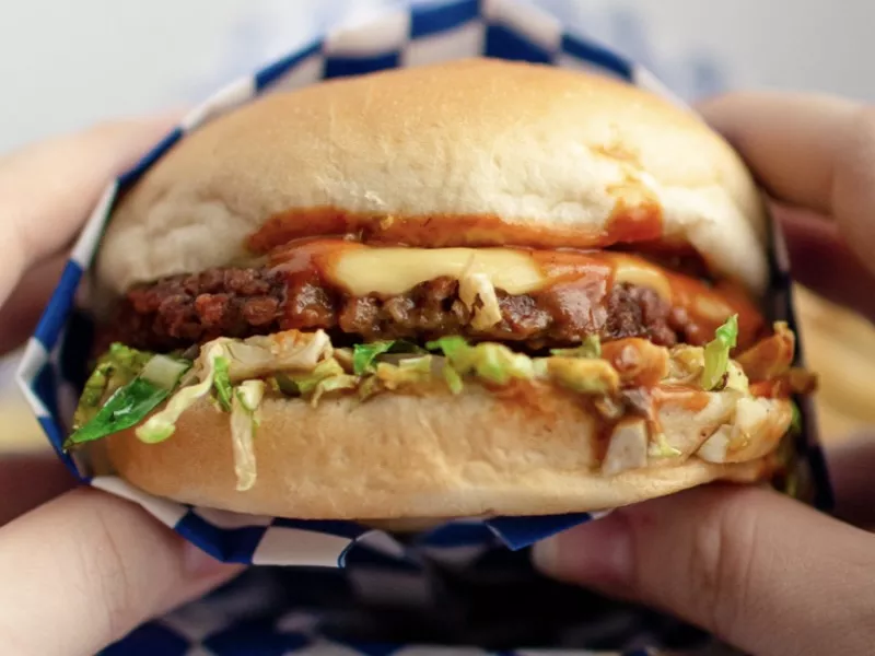 Image: Two hands holding a Meta Burger sandwich featuring a plant-based patty and vegan cheese