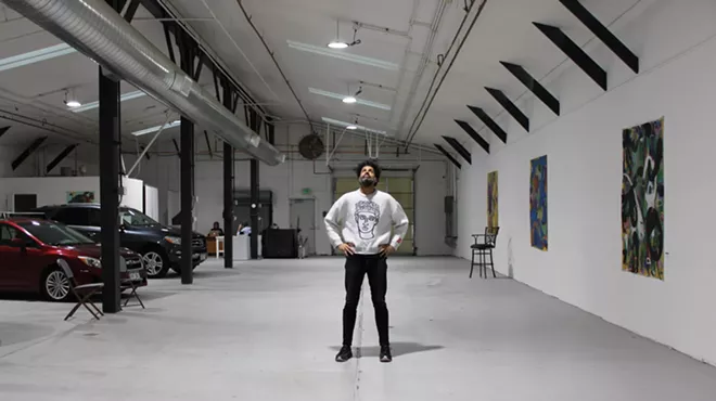 A Black man stands in the center of an art gallery/auto dealership.