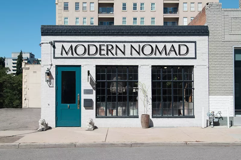 Image: The front-facing exterior of the Modern Nomad. Square building with white walls, two large windows and a cyan door. Name in large capital lettering near the roof.