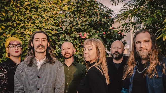 The six current members of indie rock band Murder By Death stand under some greenery.