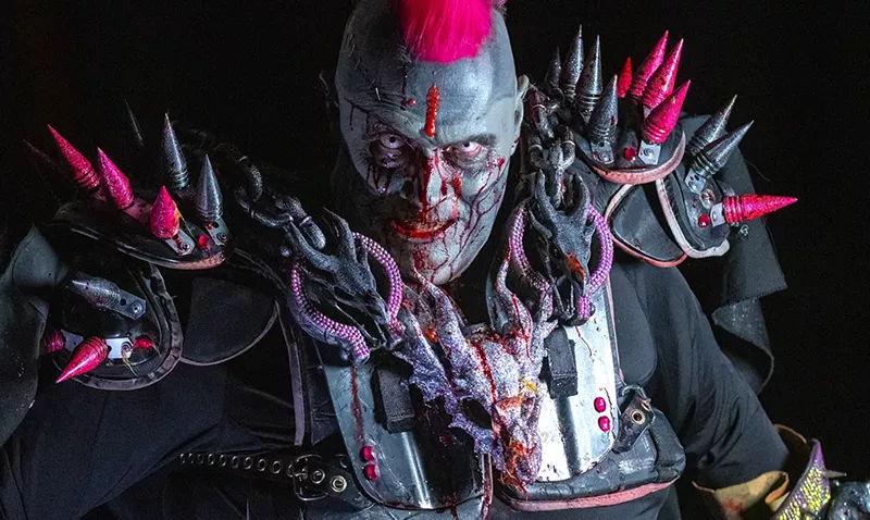 Image: man in zombie facepaint with fake blood has a pink mohawk and is wearing spiky armor