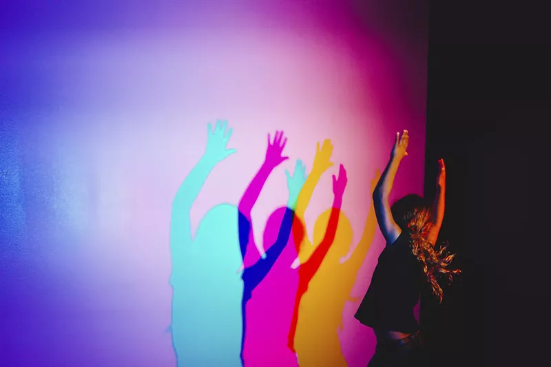 Image: a person makes colorful shadows in pink and blue on a purple wall.