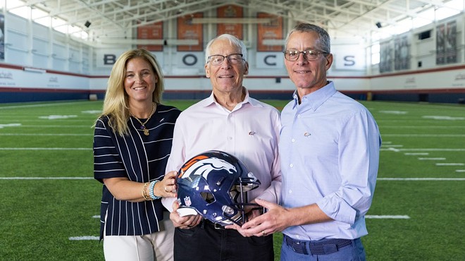 Two men and a woman hold a blue football helmet between them, smiling at the camera.