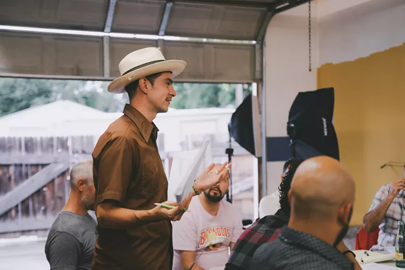 Image: man in white hat and brown shirt speaks to other seated people