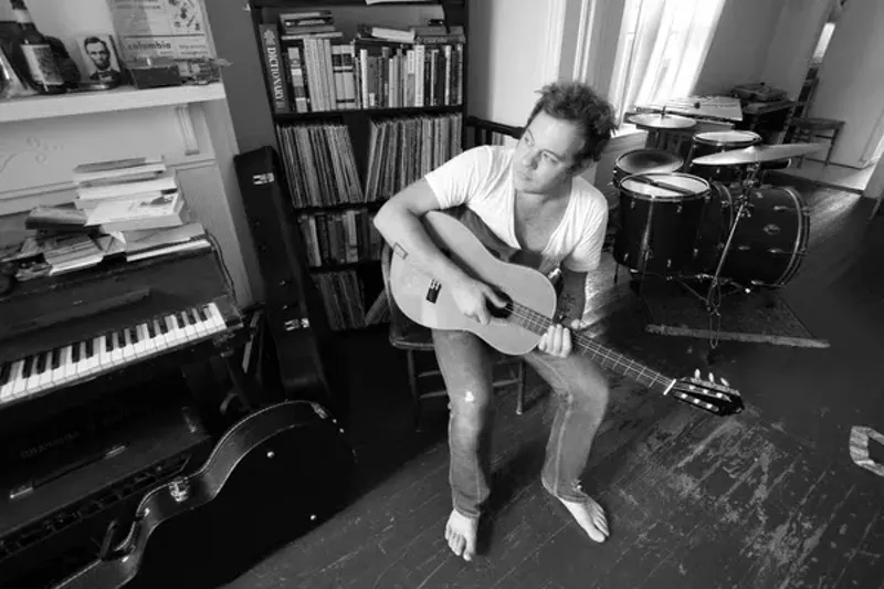 Image: a black and white photo of a man sitting strumming his acoustic guitar