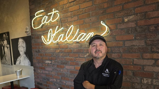 man in a black chef's coat posting in front of a neon "eat Italian" sign