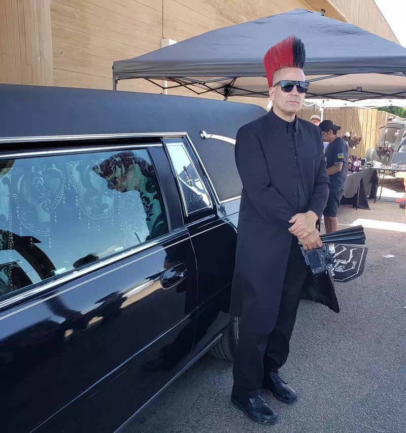 Image: A man with a mohawk stands next to a hearse