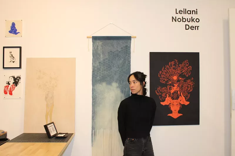 Image: A woman poses in her studio in front of her artwork.