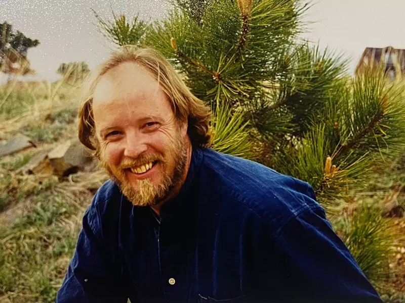 Image: A man in a blue button up smiles at the camera.