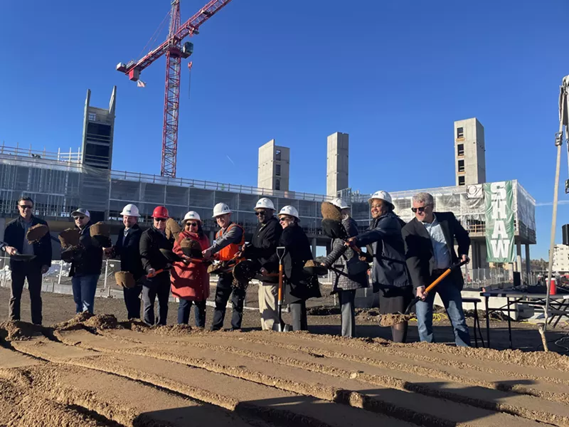 Image: A line of people wearing white hard hats toss dirt in the air using orange-handled shovels.