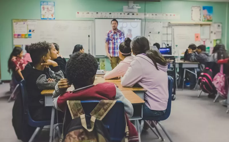 Image: Children sit at desks in a classroom, looking at an adult male teacher standing in front of a whiteboard.