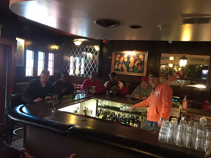 Image: a man in an orange hoodie stands behind a bar where patrons are seated