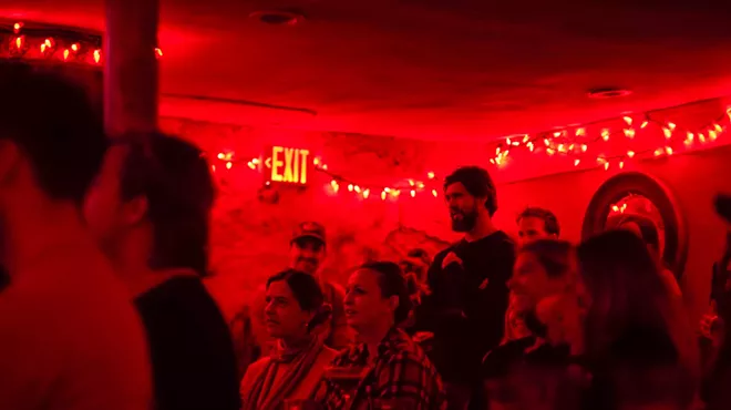 The audience sits and stands in a room lit with red lights.
