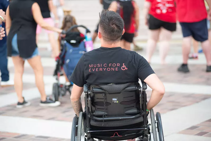 Image: Man in a wheelchair facing away with blurred bodies in front of him.