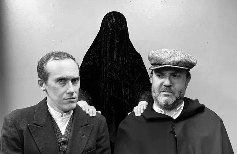 Image: a black and white photo of two men in old-fashioned clothing with a figure covered in a black drape behind them