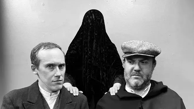 a black and white photo of two men in old-fashioned clothing with a figure covered in a black drape behind them