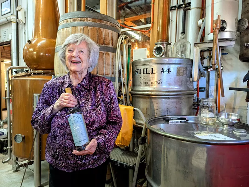 Image: woman holding a bottle of Eleanor Gin while backdropped by equipment at Spirit Hound's distillery