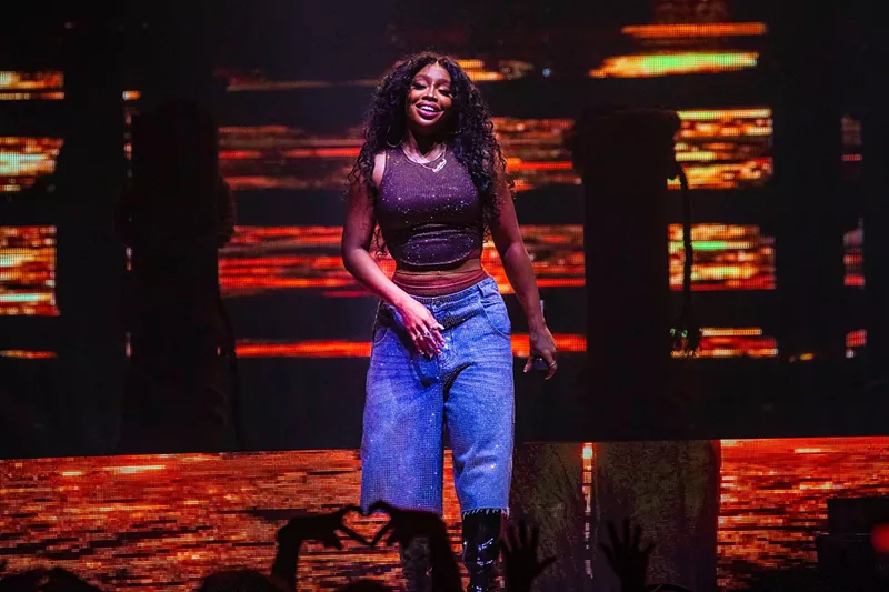 Image: R&B singer SZA in blue pants on stage with an orange background
