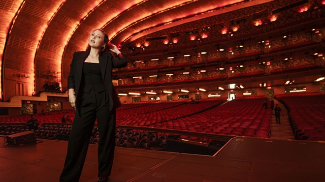 comedian taylor tomlinson stands in a theater in a black pant suit.