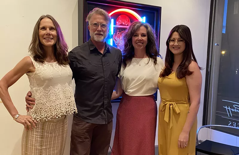 Image: woman in white shirt, man in navy shirt, woman in white shirt and skirt, and woman in yellow post in front of a painting.